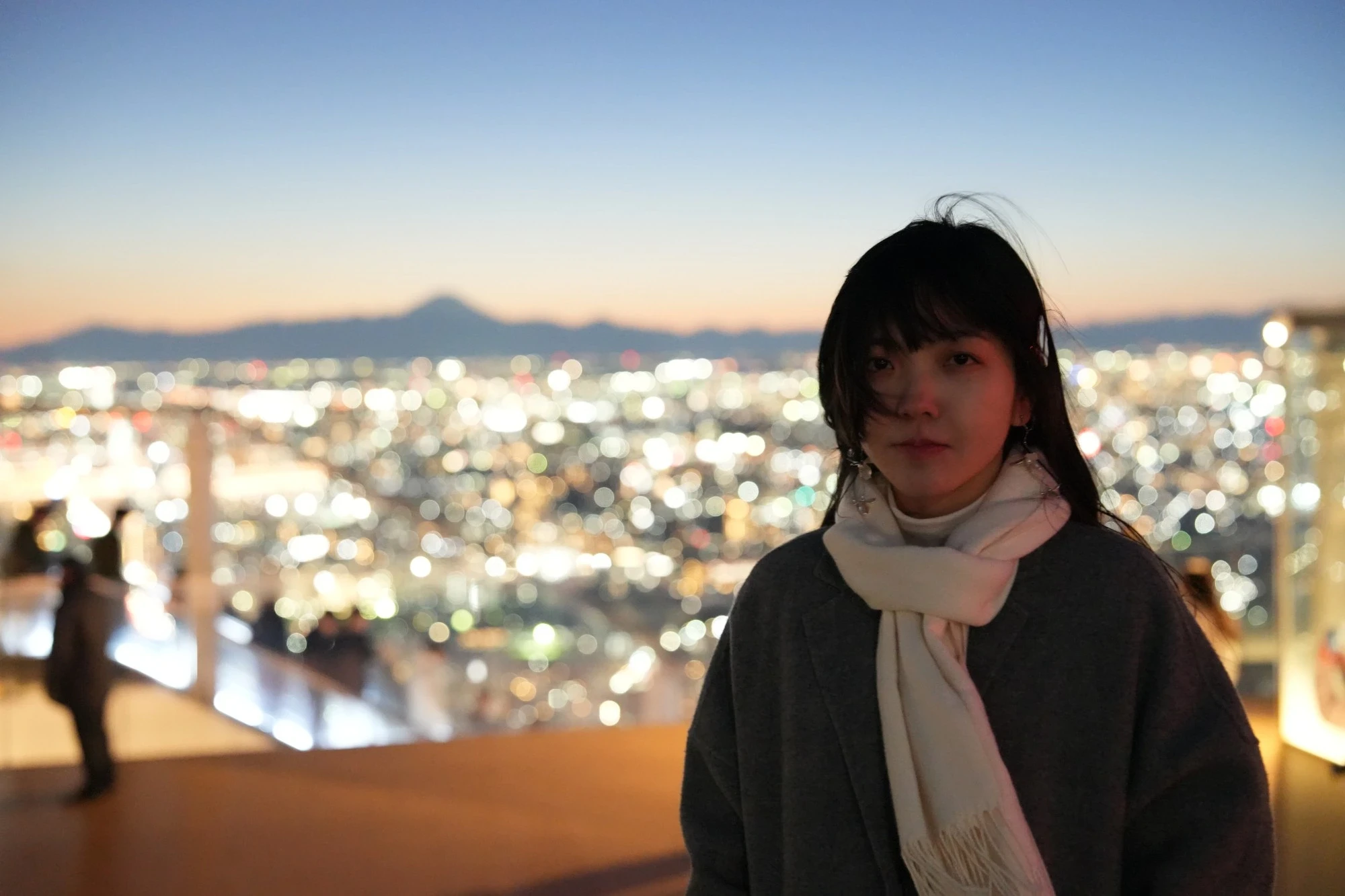 Yuki on a rooftop observation deck at dusk, in a grey coat and a white scarf, with the city lights and Mount Fuji blurred behind.
